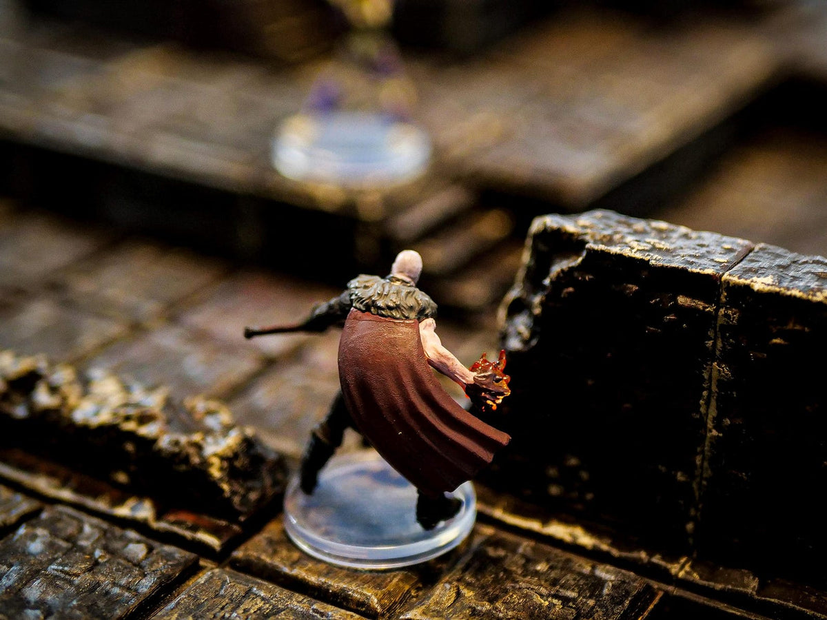 A miniature figurine of a bald character wearing a dark brown cape with a fur collar, standing on a clear circular base. The character has one arm extended, holding a small orange flame effect, and is positioned near a dark stone wall.