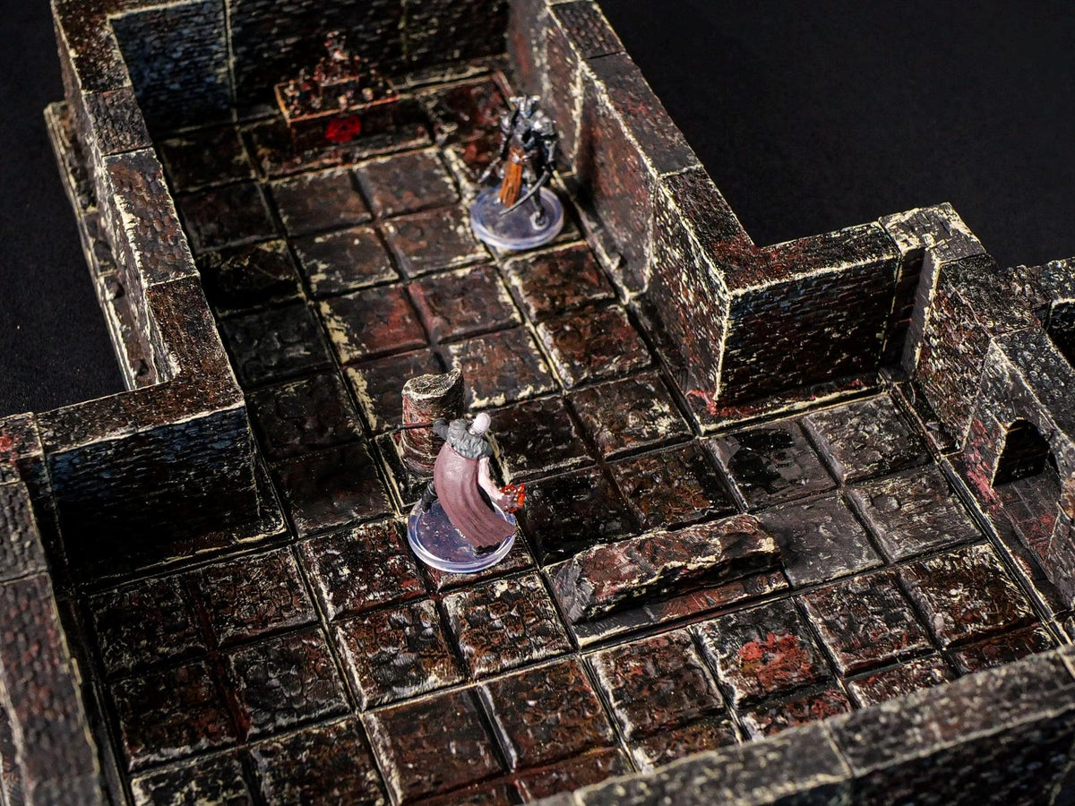 A miniature tabletop dungeon terrain set featuring dark, weathered stone walls and a grid-patterned floor with distressed brown and black textures. Two small character figurines stand on the tiles, one with a cape and one in armor.