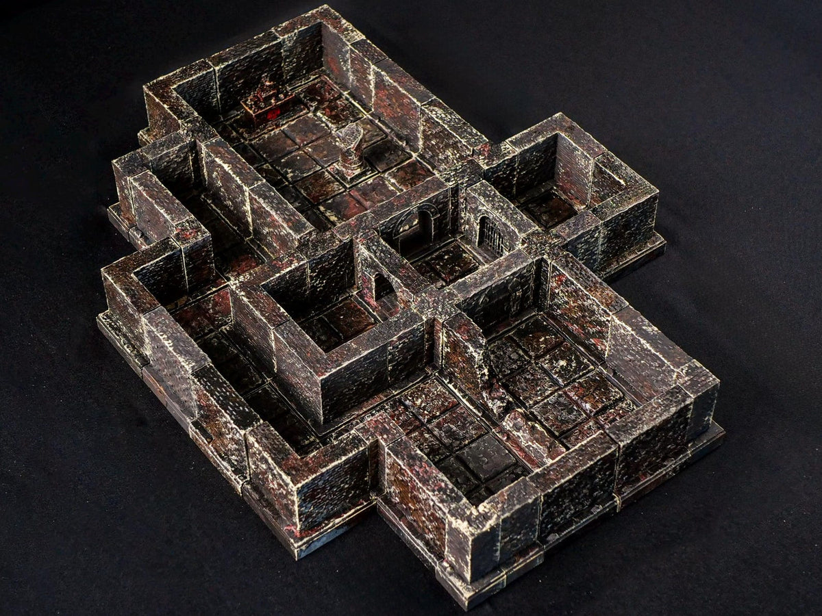 A miniature dungeon terrain set featuring interconnected stone rooms and corridors. The structure is painted in dark grey and reddish-brown tones with a weathered, textured finish, including square floor tiles and raised wall segments.