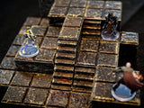 A modular tabletop gaming terrain set featuring dark, weathered stone-textured tiles arranged in a multi-level layout with stairs. Three miniature figures stand on the grid, including a skeleton, a dark armored knight, and a cloaked figure.