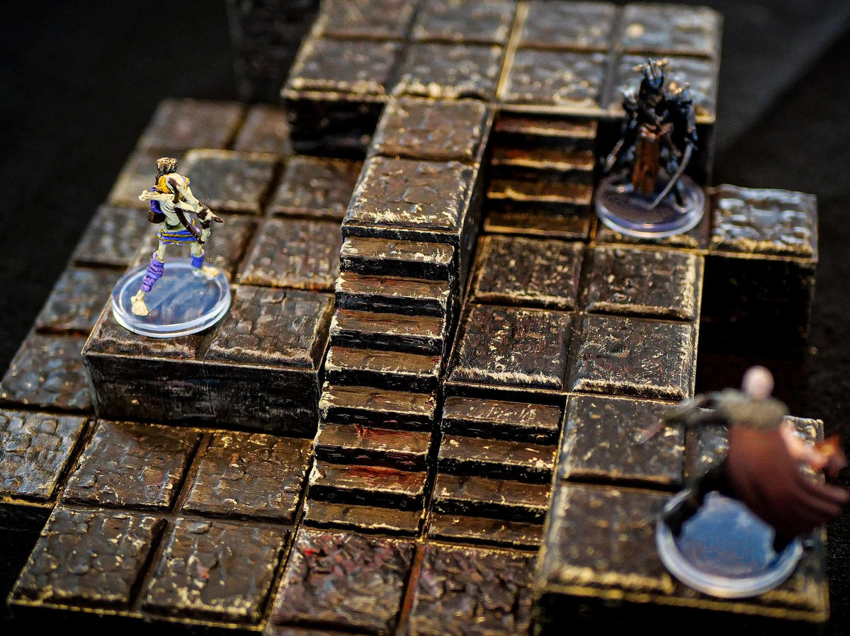 A modular tabletop gaming terrain set featuring dark, weathered stone-textured tiles arranged in a multi-level layout with stairs. Three miniature figures stand on the grid, including a skeleton, a dark armored knight, and a cloaked figure.
