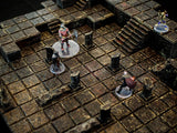 A tabletop gaming terrain set featuring a dark, weathered stone dungeon floor with a grid pattern, broken pillars, and a set of stairs. Four miniature figures stand on clear circular bases, positioned across the textured, multi-level dungeon tiles.
