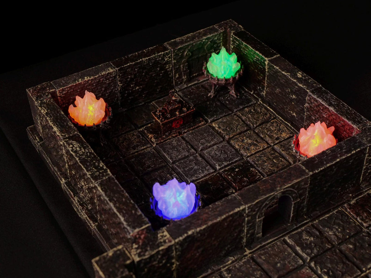 A miniature dungeon room constructed from dark, weathered stone blocks with a grid floor. The scene features four small braziers emitting glowing orange, green, and purple light, and a central rectangular chest with dark metallic details.