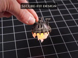 A small, metallic-colored, dome-shaped object with a dark, weathered finish and a glowing, translucent orange flame-like base. The object features a textured, architectural top and includes the text SECURE-FIT DESIGN.