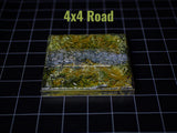 A square terrain tile featuring a central gray stone road flanked by textured green and brown grassy areas. The piece is labeled 4x4 Road in green text at the top. The surface has a detailed, uneven finish with realistic color variations.