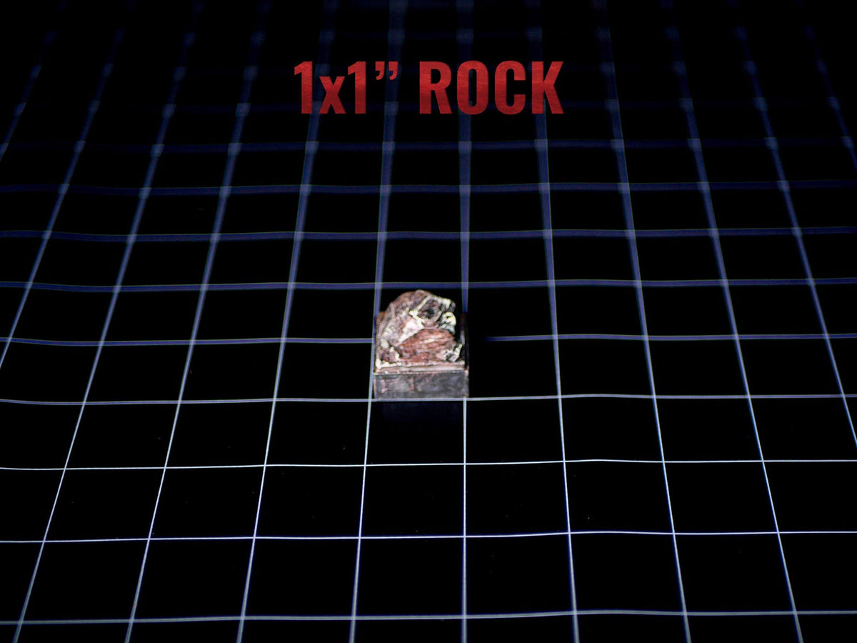 A small, textured rock with reddish-brown and grey tones, shaped into a cube. The text 1x1" ROCK is printed in red above the object, indicating its dimensions of 1 inch by 1 inch. The rock has a rough, natural surface texture.