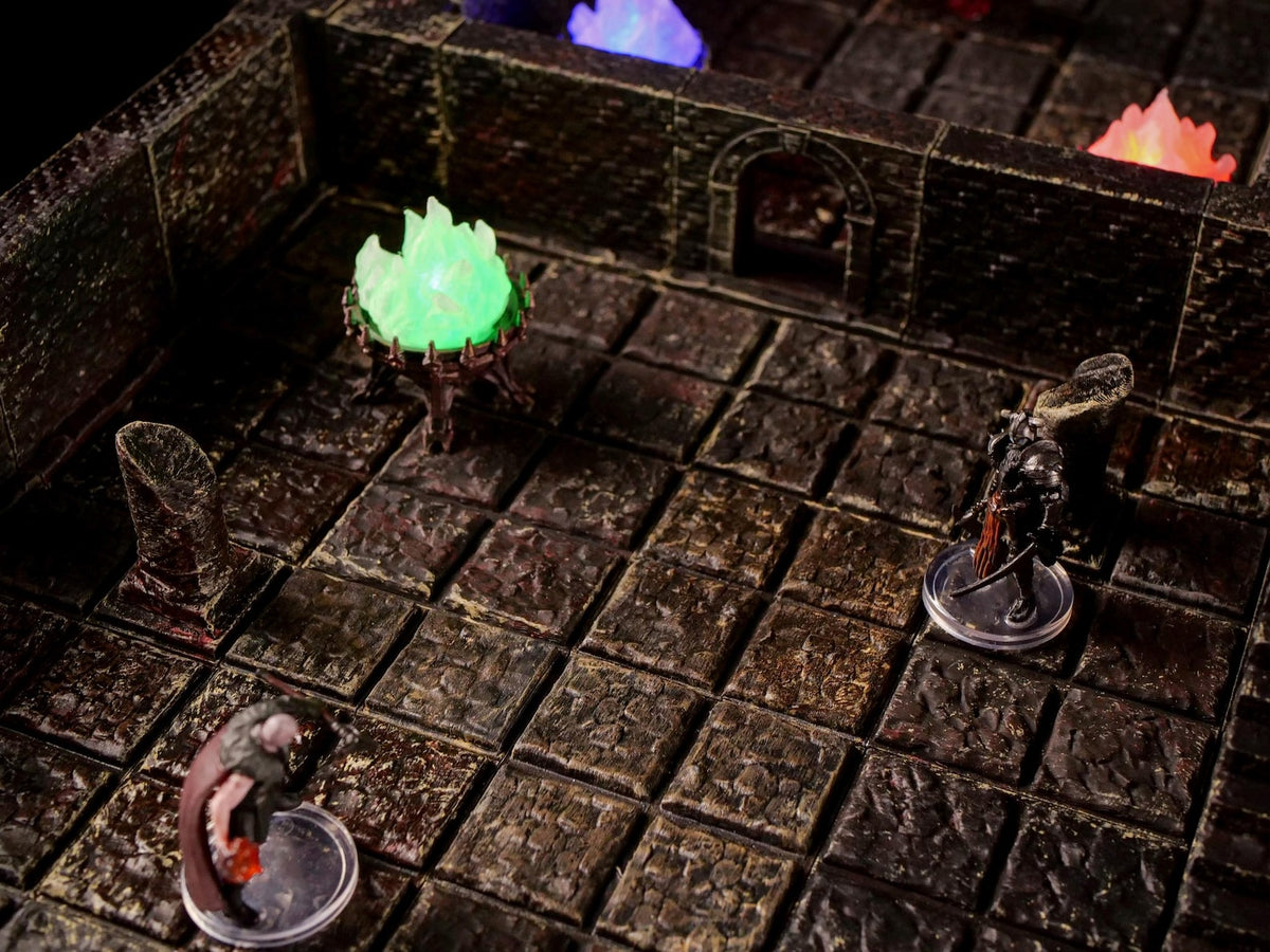 A tabletop gaming dungeon terrain set featuring a dark stone grid floor with textured walls, a glowing green fire brazier, and two miniature figurines standing on clear circular bases within the modular stone room environment.