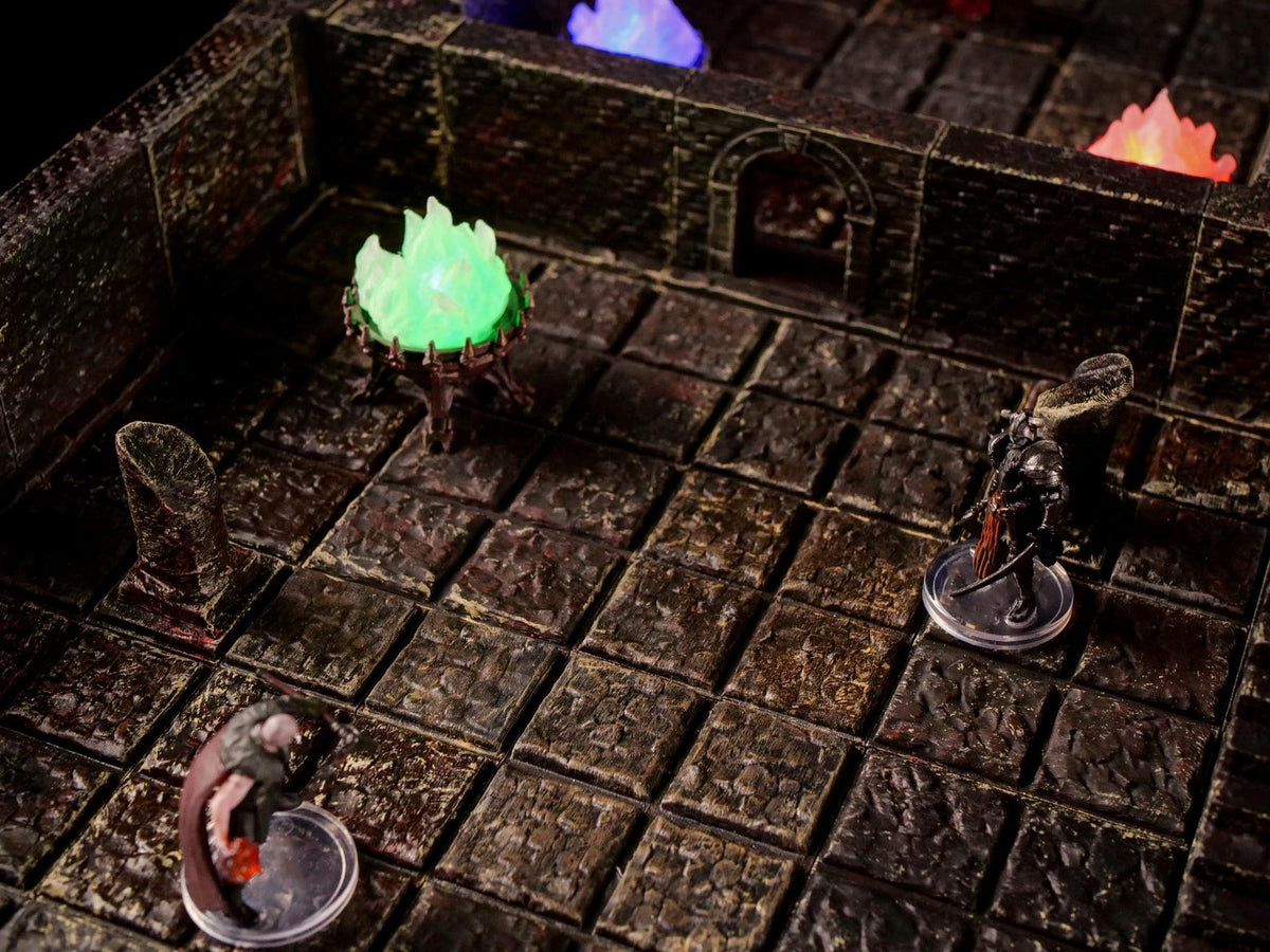 A tabletop gaming dungeon terrain set featuring a dark stone grid floor with textured walls, a glowing green fire brazier, and two miniature figurines standing on clear circular bases within the modular stone room environment.