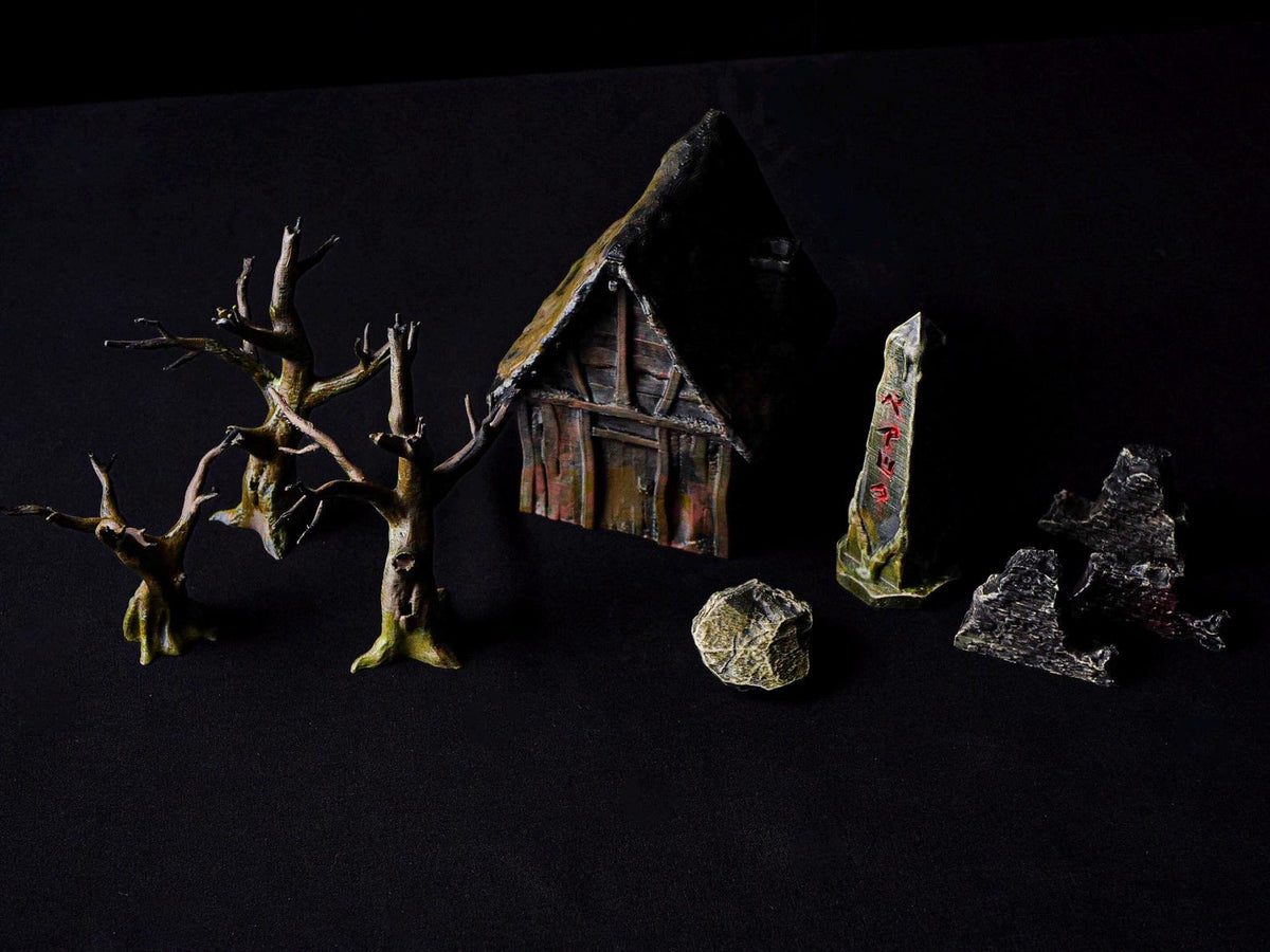 A miniature tabletop gaming set featuring a small wooden-style cottage with a dark roof, three leafless brown trees, a grey stone with red runic markings, a textured rock, and two jagged grey stone fragments on a black background.