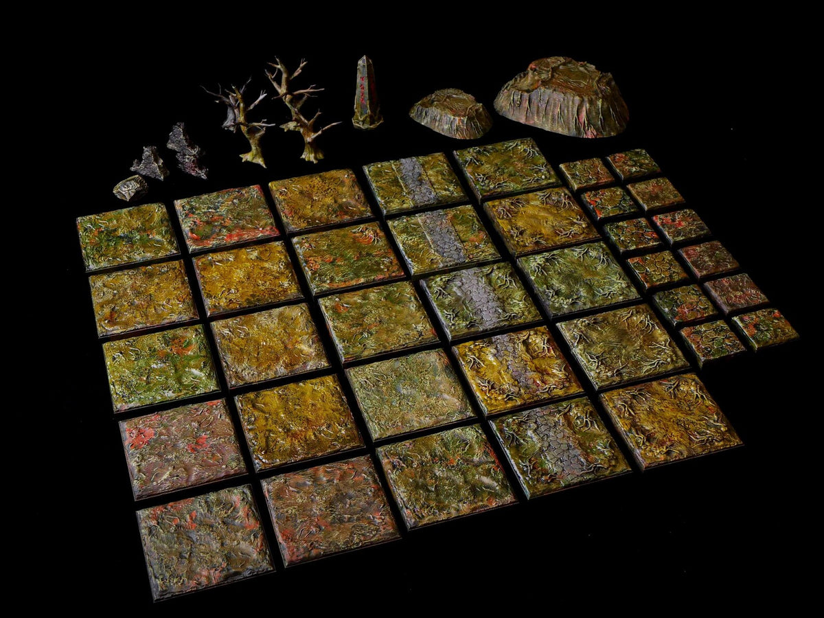 A collection of miniature tabletop gaming terrain pieces, including a grid of square tiles with textured green and brown forest floor patterns, two small leafless trees, a stone obelisk, and two rocky outcroppings on a black surface.