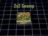A square game tile featuring a textured swamp landscape with shades of green, brown, and yellow paint. The top surface displays a detailed terrain pattern, and the text 2x2 Swamp is printed in light green above the object.