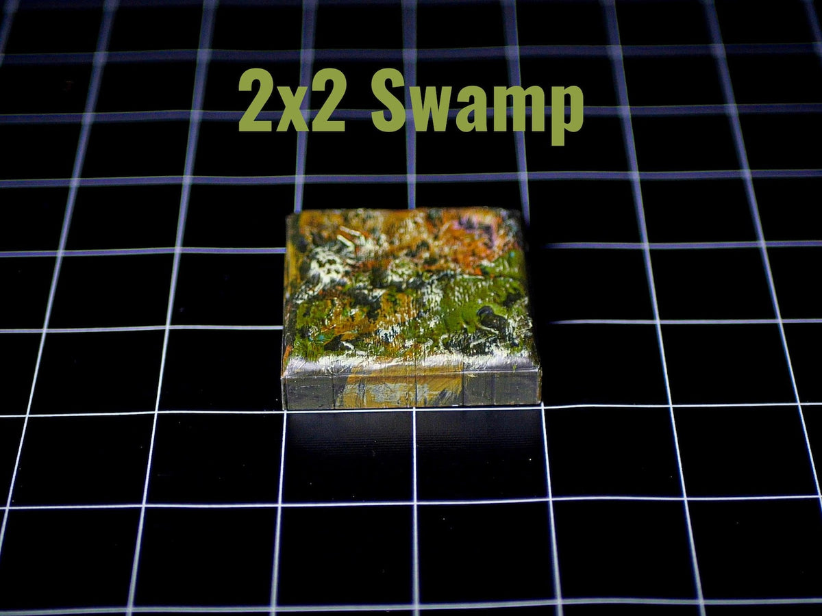 A square game tile featuring a textured swamp landscape with shades of green, brown, and yellow paint. The top surface displays a detailed terrain pattern, and the text 2x2 Swamp is printed in light green above the object.