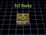 A square tile with a textured, multi-colored surface featuring shades of green, brown, and orange, resembling a rocky terrain. The text 2x2 Rocky is printed in light green above the tile, which is set against a black grid background.