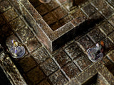 A miniature tabletop gaming terrain set featuring a dark brown stone dungeon floor with a grid pattern and high walls. Two small painted figurines stand on clear circular bases within the corridors of the modular stone structure.
