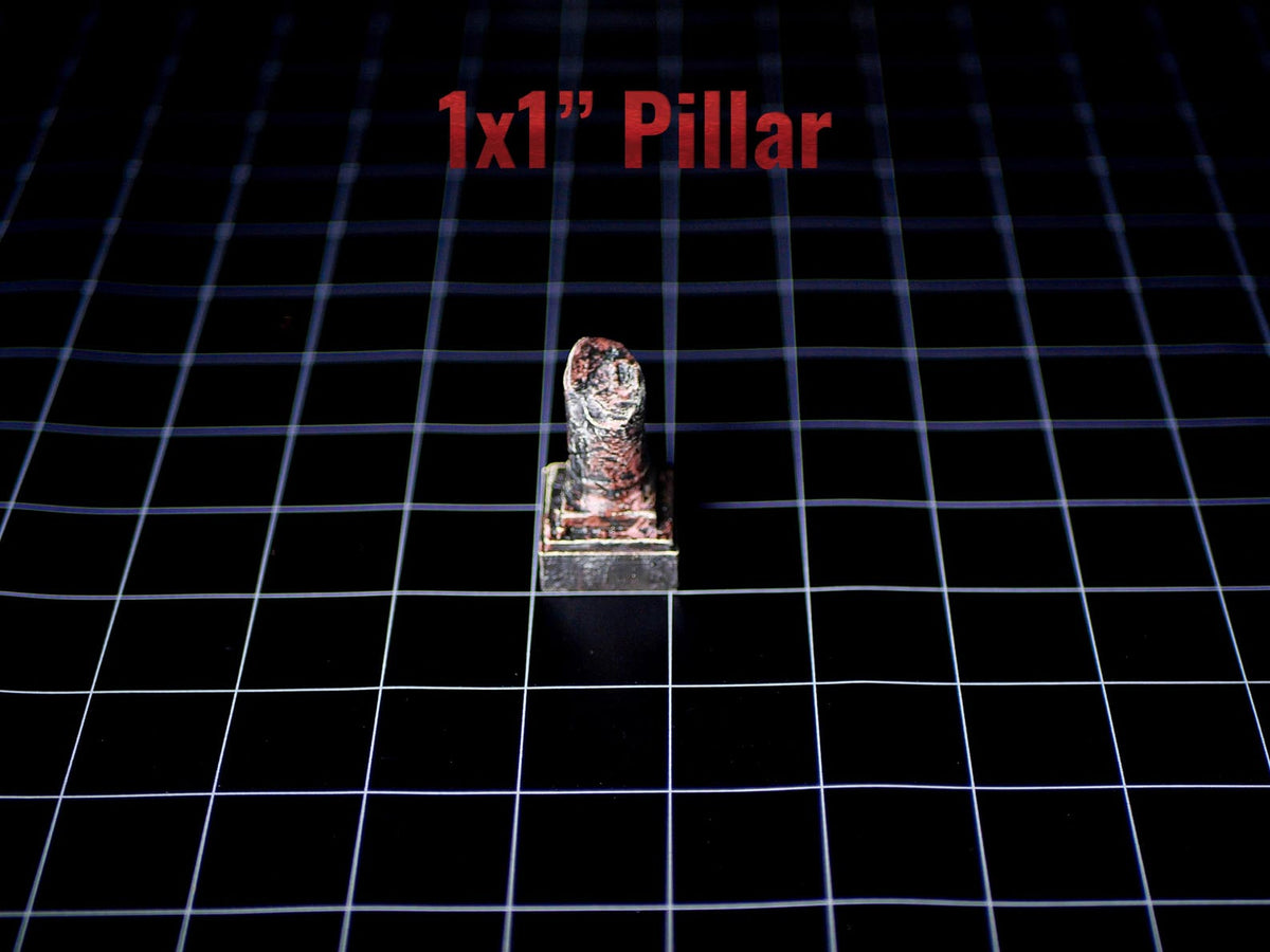 A miniature 1x1 inch pillar with a weathered, dark, and reddish-brown textured finish. The object features a square base and a cylindrical top section, presented on a black grid surface with the text 1x1 inch Pillar visible.