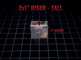 A rectangular riser block with a dark, weathered finish featuring patches of reddish-brown texture. The top surface has a narrow slit. Text on the image reads 2x1" RISER - TALL and 2" HEIGHT, indicating a height of 2 inches.