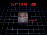A square 2x2 inch riser with a 1 inch height, featuring a textured, weathered metallic finish with dark grey and rust-colored patches. The top surface is divided into four smaller squares, and the text 2x2 RISER - MID is visible.