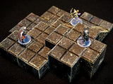 A modular dungeon terrain set composed of dark brown, stone-textured square blocks arranged in a stepped layout. Three small painted fantasy character figurines stand on clear circular bases atop the grid-patterned terrain tiles.