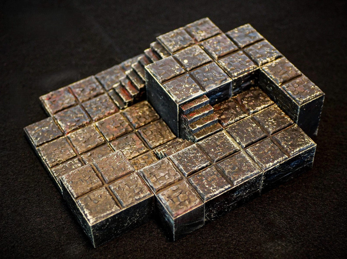 A modular tabletop gaming terrain piece featuring a multi-level stone floor design with integrated stairs. The structure is composed of dark, weathered square tiles with a textured, metallic finish and visible bronze-toned highlights.