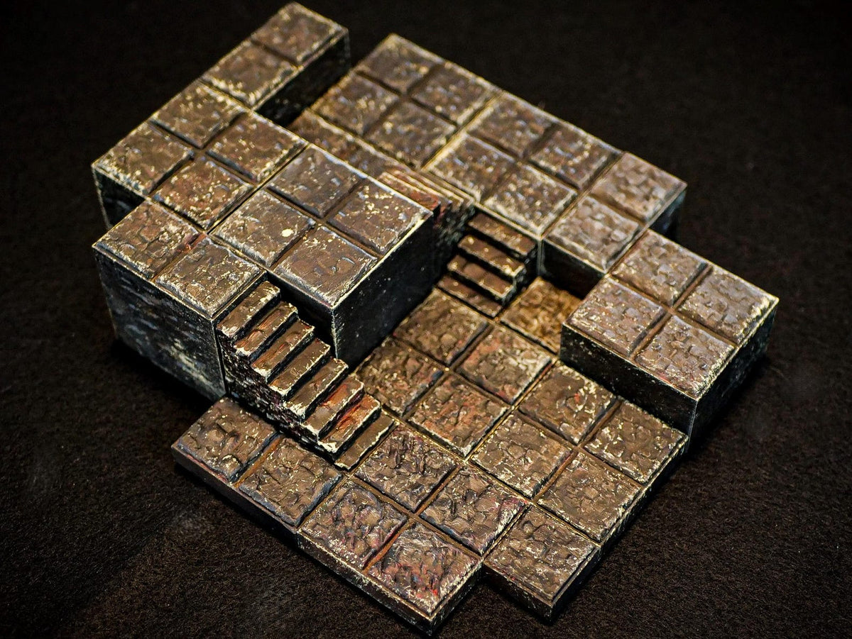 A miniature dungeon terrain piece featuring a multi-level stone floor layout with raised platforms and a set of stairs. The surface is textured with a dark brown and grey weathered finish, resembling aged stone tiles in a grid pattern.