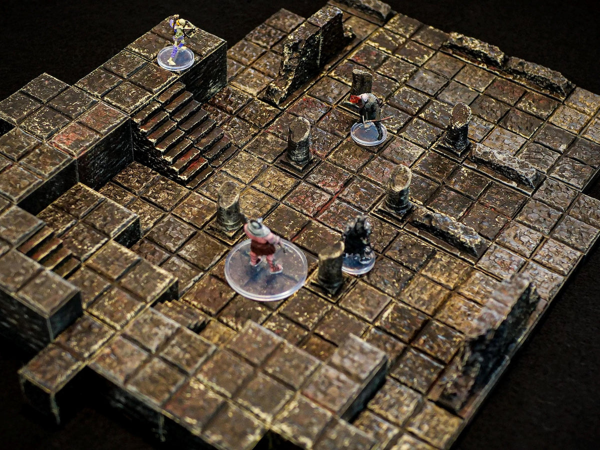 A modular dungeon tile set featuring a grid of weathered, dark brown stone floor tiles with raised walls, broken pillars, and stone staircases. Four small painted miniature figures stand on clear circular bases across the terrain.