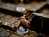A miniature figurine of a bald character wearing a dark brown cape with a fur collar, standing on a clear circular base. The character has one arm extended, holding a small orange flame effect, and is positioned near a dark stone wall.