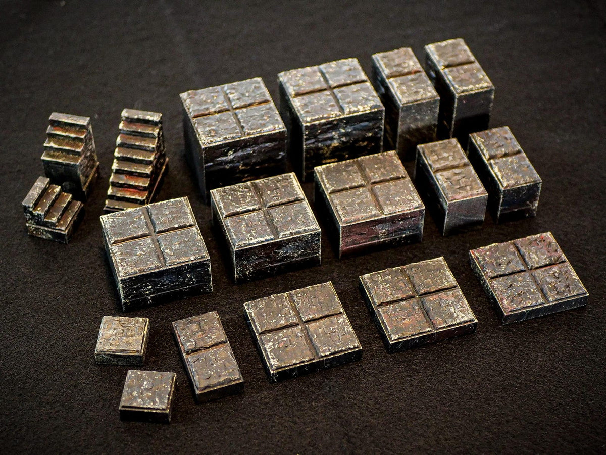 A collection of dark, metallic-toned miniature blocks and staircases with a weathered, textured surface. The set includes various square and rectangular pieces, some featuring a cross-shaped indentation on the top surface.