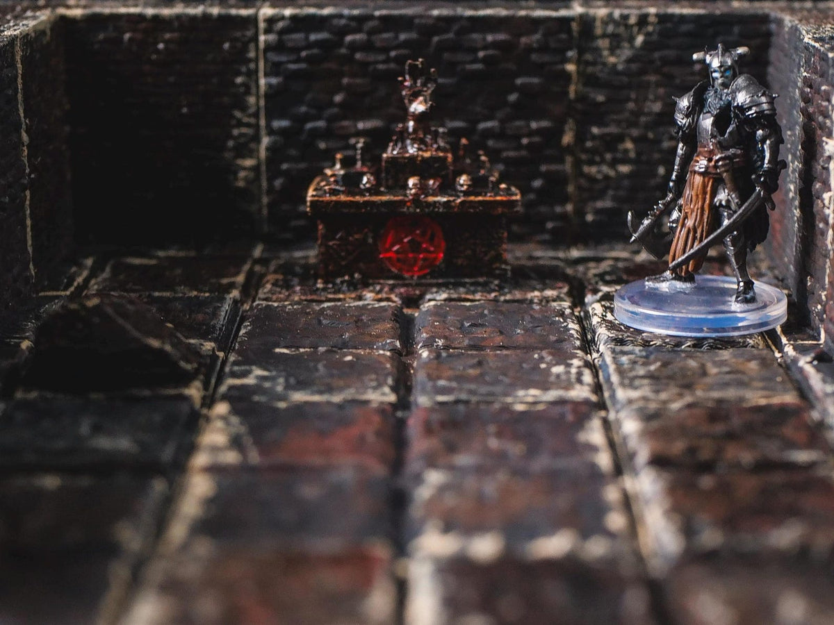A miniature armored warrior figure stands on a circular base within a dark stone dungeon setting. Nearby, a small altar features a red pentagram symbol on its front, with skulls and a flame-like structure resting on its top surface.
