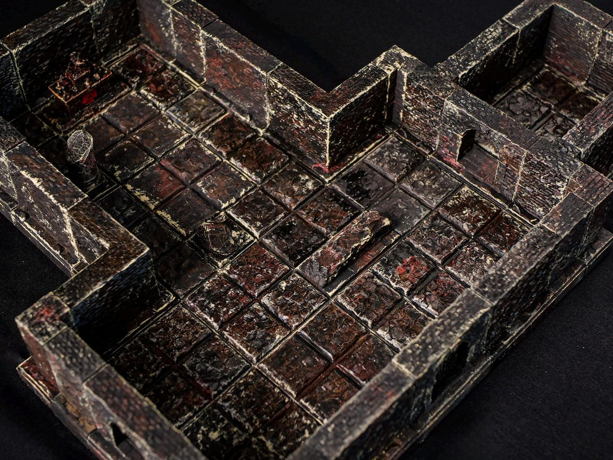 A miniature tabletop dungeon terrain piece featuring a dark, weathered stone floor with a grid pattern. The walls are textured with a distressed black and dark red finish, including a small altar in the corner and a broken pillar.
