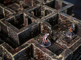 A modular dungeon terrain set featuring dark, weathered stone walls with a textured finish. Two miniature figures stand on the grid floor, one wearing a dark cloak and the other in silver armor, positioned within the stone corridors.