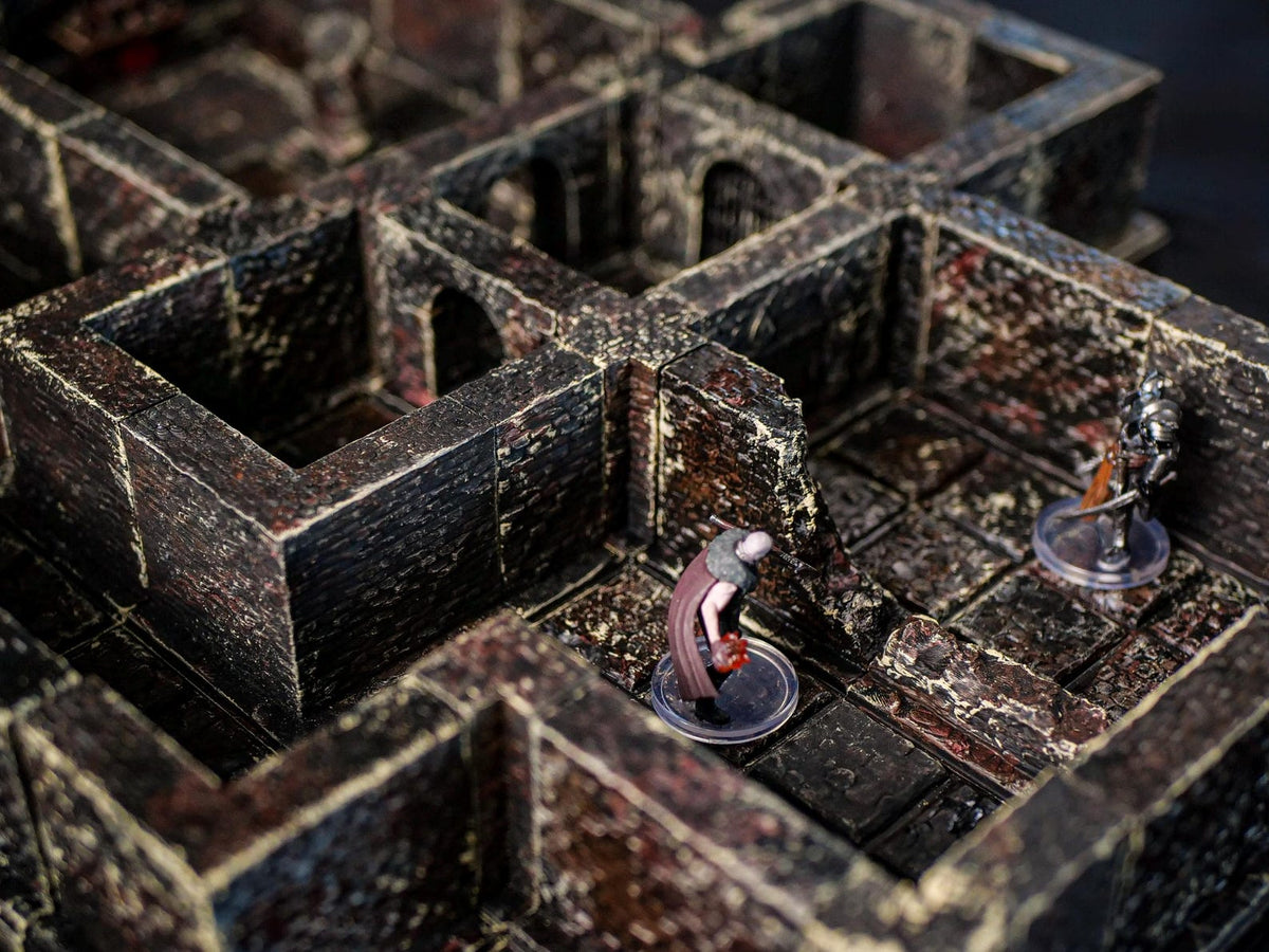 A modular dungeon terrain set featuring dark, weathered stone walls with a textured finish. Two miniature figures stand on the grid floor, one wearing a dark cloak and the other in silver armor, positioned within the stone corridors.