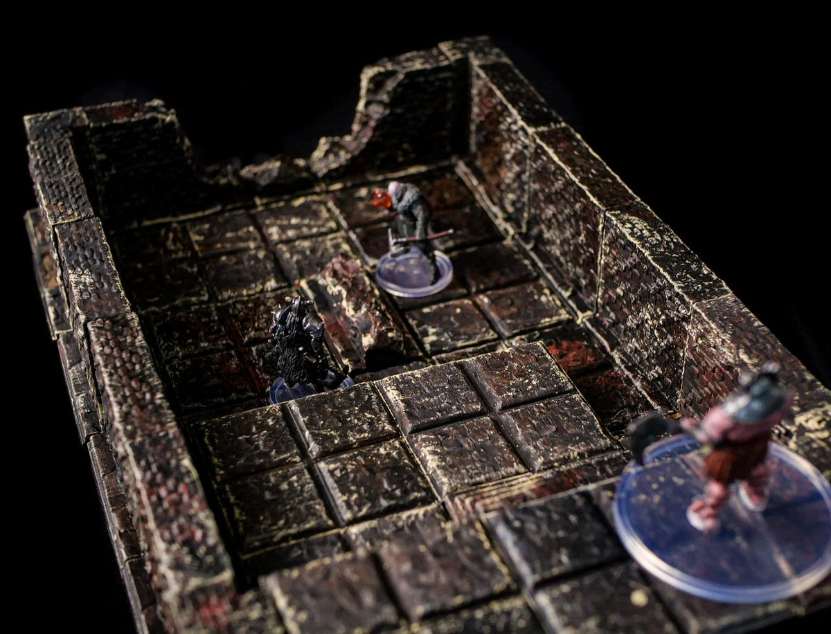 A detailed, miniature dungeon terrain set with a textured, stone-like appearance. The set features a grid floor, walls, and several miniature figures on clear bases, creating a scene for tabletop gaming.