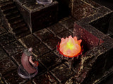 A miniature tabletop gaming scene featuring a dark stone dungeon floor with a staircase and a wall. A small figure in a maroon cloak stands near a dark metal brazier containing a bright, translucent orange and yellow flame effect.