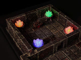 A miniature dungeon room constructed from dark, weathered stone blocks with a grid floor. The scene features four small braziers emitting glowing orange, green, and purple light, and a central rectangular chest with dark metallic details.