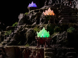 Three decorative braziers with glowing flames in green, orange, and purple, positioned on a dark, rocky, tiered landscape. Each metal-toned bowl features a spiked rim and holds a translucent, light-emitting flame sculpture.