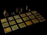 A collection of miniature tabletop gaming terrain pieces including a small house, two bare trees, a stone obelisk, and various square tiles featuring textured green, brown, and yellow forest floor patterns with scattered debris.