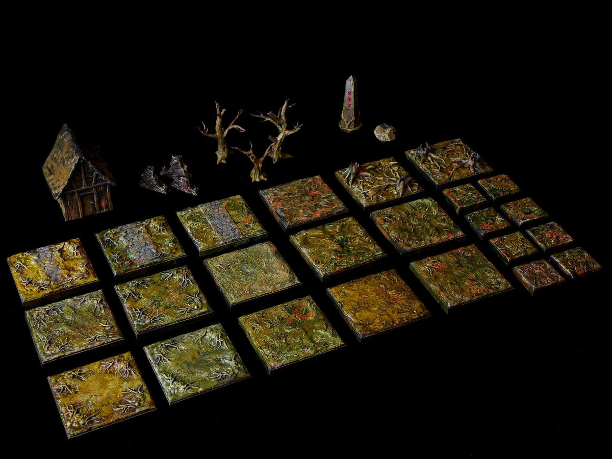 A collection of miniature tabletop gaming terrain pieces including a small house, two bare trees, a stone obelisk, and various square tiles featuring textured green, brown, and yellow forest floor patterns with scattered debris.