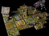 A tabletop gaming terrain set featuring modular square tiles with a swampy, overgrown texture in shades of green and brown. The set includes a small house, a central stone obelisk with red markings, bare trees, rocks, and a cloth dice bag.