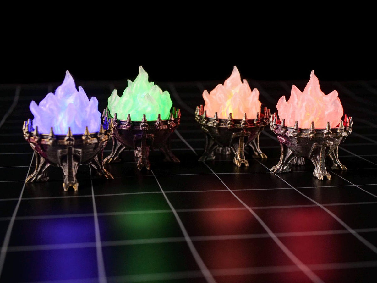 Four miniature metal-colored braziers with spiked rims, each containing a translucent flame-shaped insert. The flames are illuminated in blue, green, orange, and pink, casting a glow on the dark surface beneath the objects.