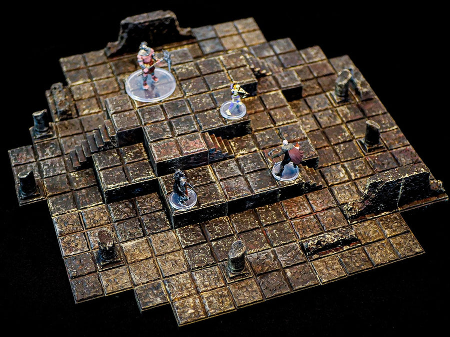 A multi-level dungeon terrain set featuring a dark, weathered stone tile grid with raised platforms, staircases, and broken pillar ruins. Four miniature figures on clear circular bases are positioned across the modular stone layout.
