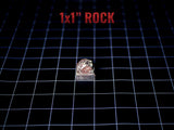 A small, textured rock with reddish-brown and grey tones, shaped into a cube. The text 1x1" ROCK is printed in red above the object, indicating its dimensions of 1 inch by 1 inch. The rock has a rough, natural surface texture.