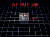 A rectangular metal riser with a weathered, dark grey finish and a central vertical groove. The object is labeled with the text 2x1" RISER - MID and features a red indicator line showing a 1" height measurement on the side.