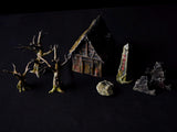 A miniature tabletop gaming set featuring a small wooden-style cottage with a dark roof, three leafless brown trees, a grey stone with red runic markings, a textured rock, and two jagged grey stone fragments on a black background.