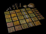 A collection of miniature tabletop gaming terrain pieces, including a grid of square tiles with textured green and brown forest floor patterns, two small leafless trees, a stone obelisk, and two rocky outcroppings on a black surface.