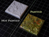Two square terrain tiles for tabletop gaming. One tile is unpainted white resin with a textured surface, labeled "NOT PAINTED". The other tile is painted with green, brown, and orange tones, labeled "PAINTED". Both have a flat base.