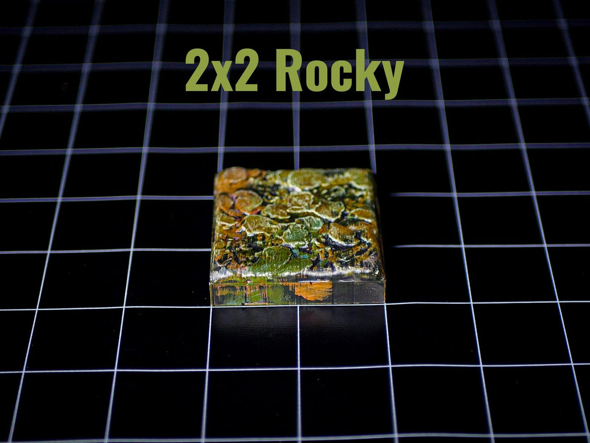 A square tile with a textured, multi-colored surface featuring shades of green, brown, and orange, resembling a rocky terrain. The text 2x2 Rocky is printed in light green above the tile, which is set against a black grid background.