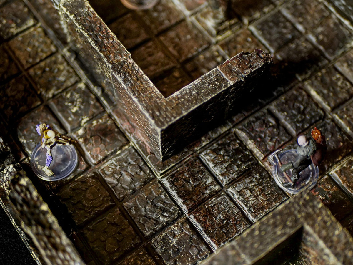A miniature tabletop gaming terrain set featuring a dark brown stone dungeon floor with a grid pattern and high walls. Two small painted figurines stand on clear circular bases within the corridors of the modular stone structure.