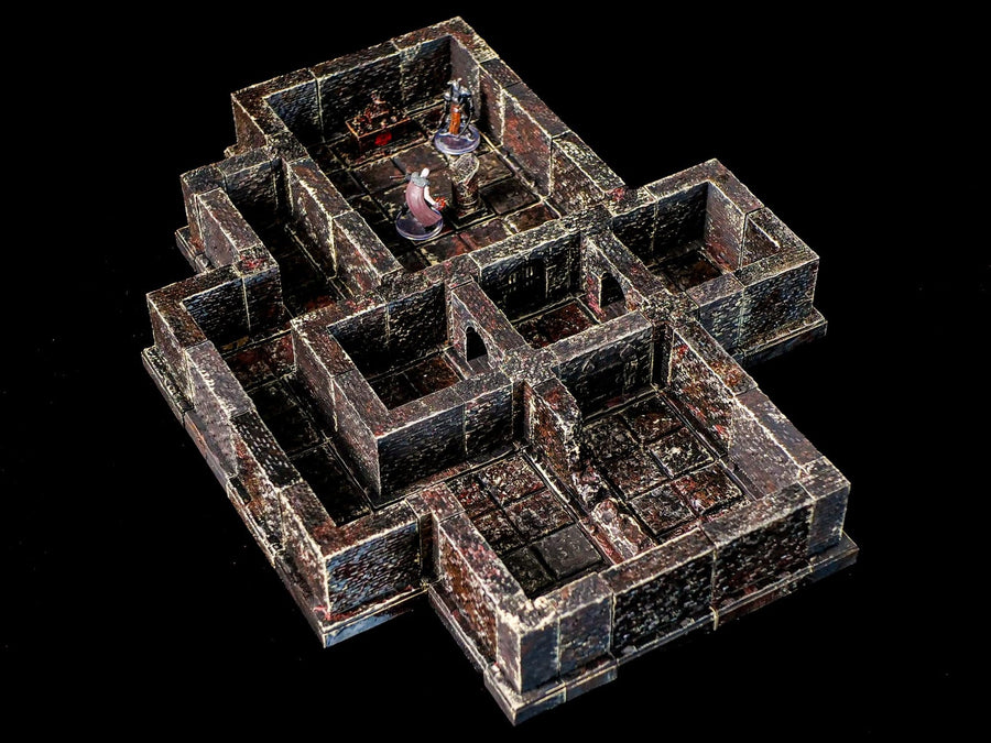 A miniature dungeon terrain set featuring dark, weathered stone walls and floor tiles with reddish-brown highlights. The modular structure includes multiple interconnected rooms and corridors, with two small humanoid figurines placed inside.