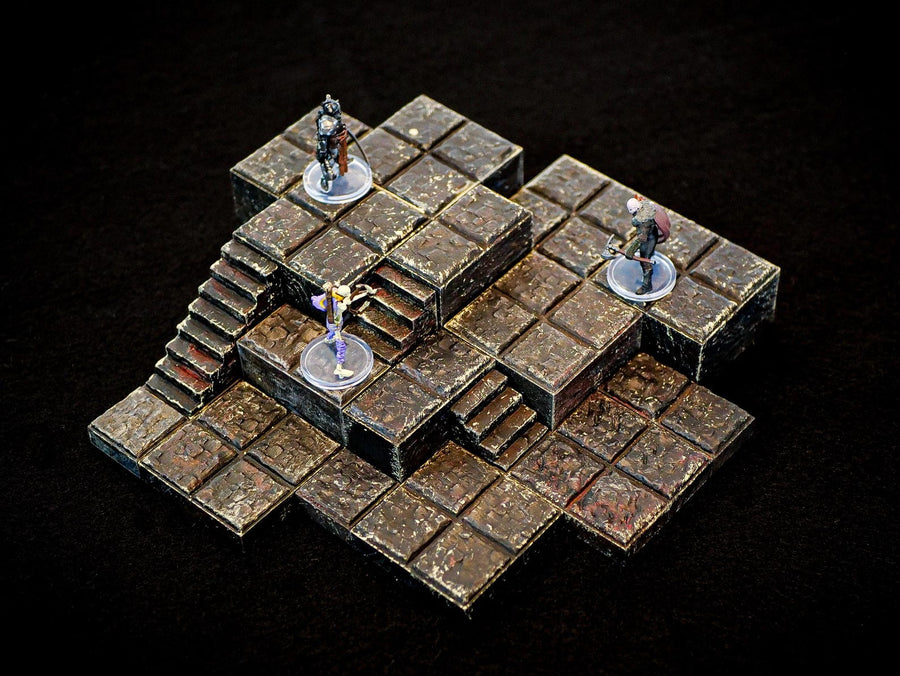 A multi-level dungeon terrain set featuring dark, weathered stone tiles with a grid pattern. The structure includes raised platforms and integrated staircases, with three small painted character miniatures standing on clear circular bases.