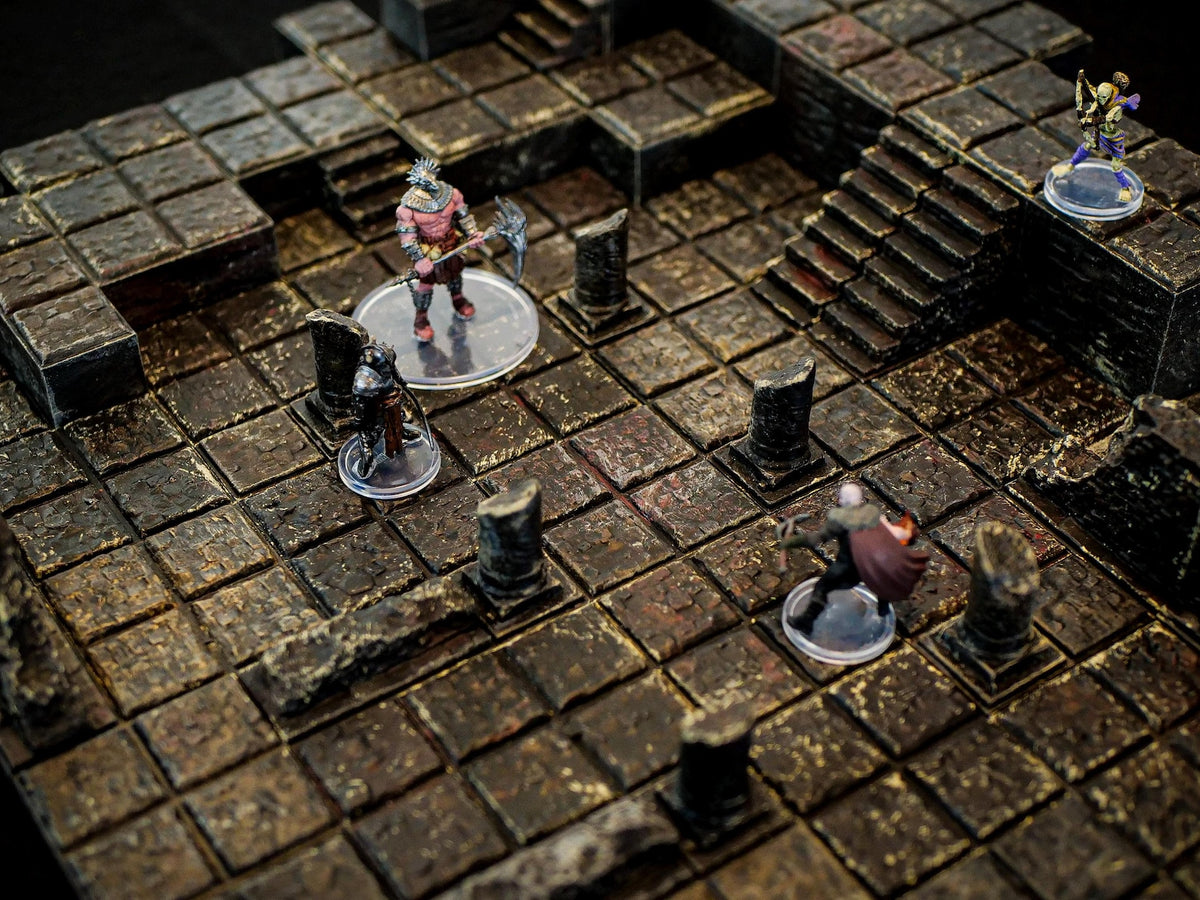 A tabletop gaming terrain set featuring a dark, weathered stone dungeon floor with a grid pattern, broken pillars, and a set of stairs. Four miniature figures are positioned on the tiles, each standing on a clear circular base.